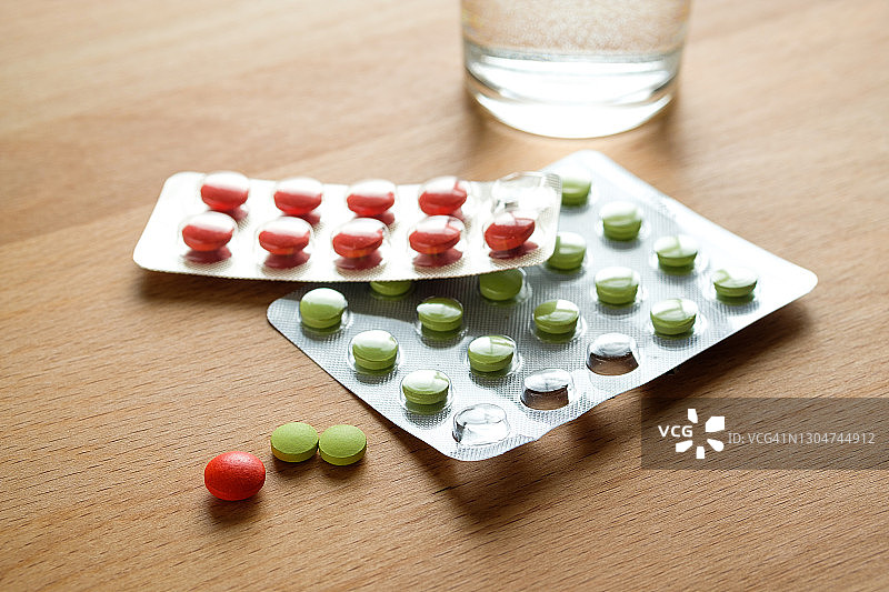 红色和绿色的药丸和一杯水放在厨房的木制桌子上。服用维生素、止痛药、药物和膳食补充剂。疾病的概念预防和治疗，保健和医药。计划和准备怀孕。图片素材