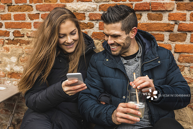 微笑的女人用智能手机，而男人拿着果汁杯靠墙坐着图片素材