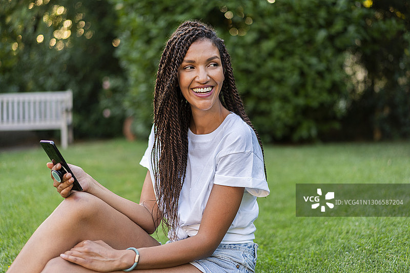 微笑的编着辫子的女人与手机看向别处，而坐在花园里在夏天图片素材