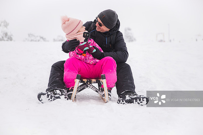 快乐的父亲和女儿拿着GoPro相机，看着对方当雪橇图片素材