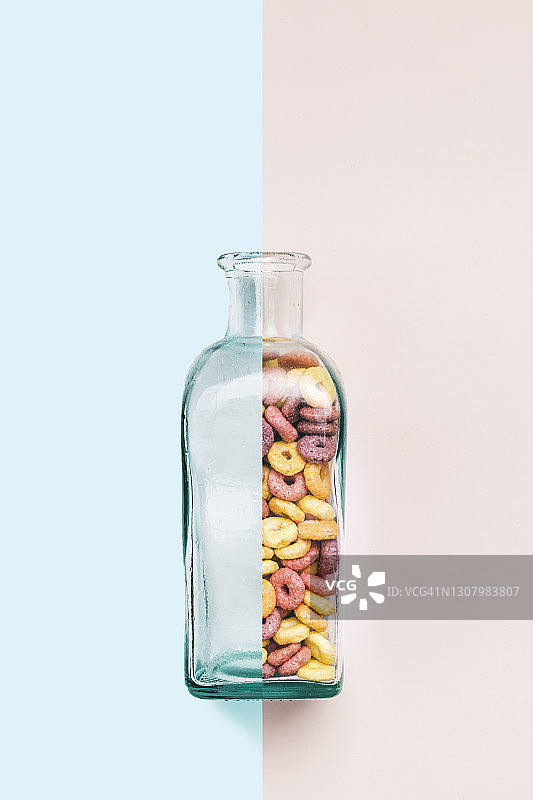 这是一个空的和满的玻璃瓶，里面有彩色的圆环形状的谷物，背景是浅粉色和蓝色。极简主义的概念图片素材