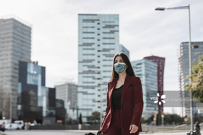 在新冠肺炎期间，身着红色西装的女商人戴着防护口罩在城市行走图片素材