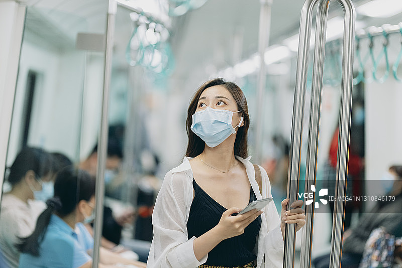 亚洲女性在乘坐地铁或火车时，会戴口罩、使用智能手机和耳机，并保持社交距离图片素材