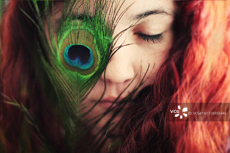 一个闭着眼睛的红头发女人的肖像。在他的一只眼睛前面有一根美丽的孔雀羽毛。图片素材