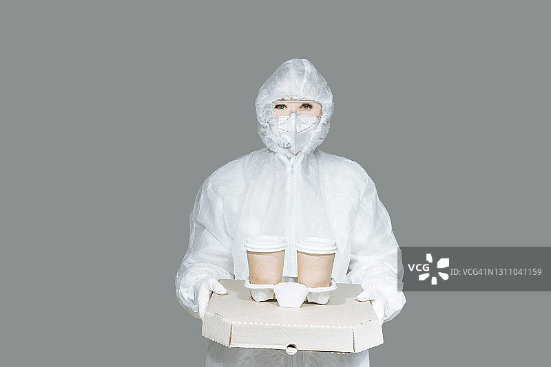 身穿防护服、戴着手套和面罩的年轻女孩在灰色背景下送食物，送咖啡和披萨。图片素材