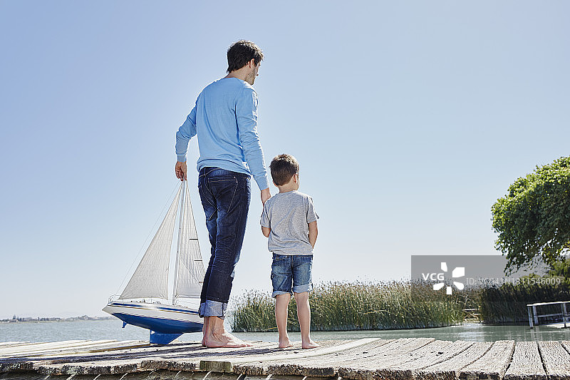 成熟的男人拿着玩具帆船牵着小男孩的手，对着天空站着图片素材