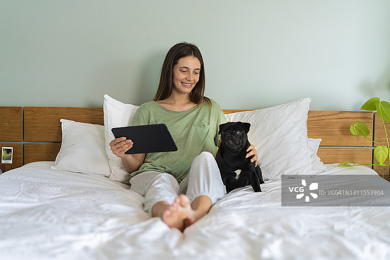 一个年轻女子拿着平板电脑，和帕格狗坐在家里的床上图片素材