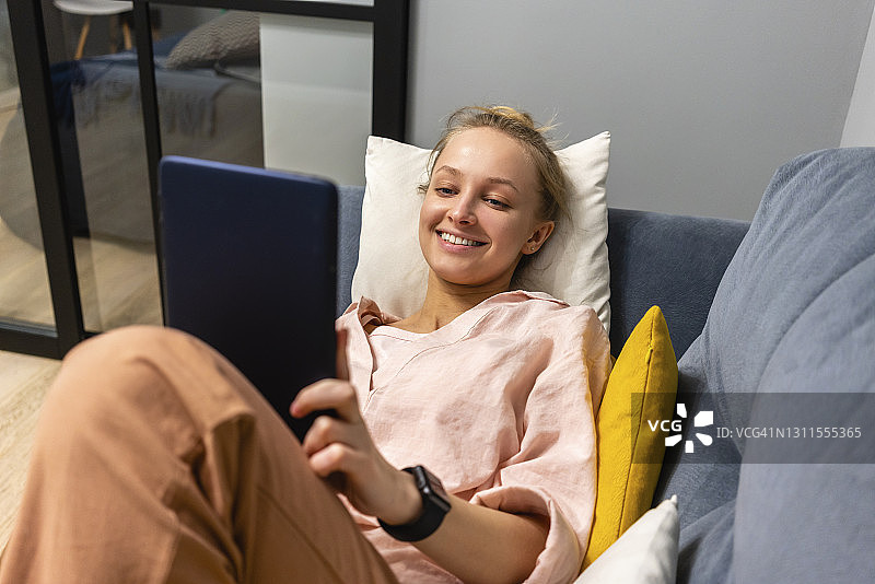 微笑的年轻女子躺在家里的沙发上使用电子平板电脑图片素材