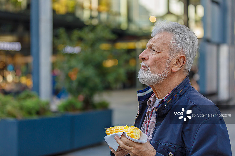 一位老人在城市里散步，享受玉米芯。图片素材