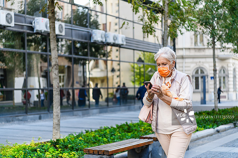 一位上了年纪的妇女在流感大流行期间匆忙地使用手机行走在城市街道上。图片素材