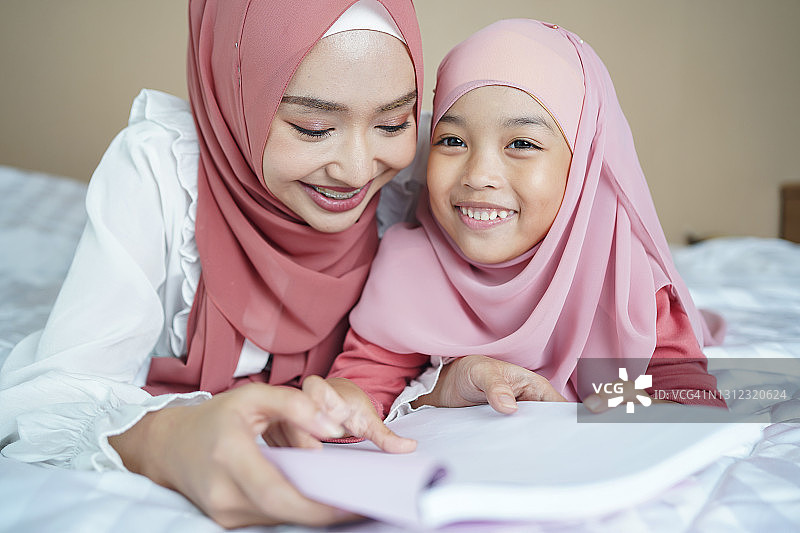 年轻的亚洲穆斯林母亲在家里的卧室里教她的小女儿阅读这本书。图片素材
