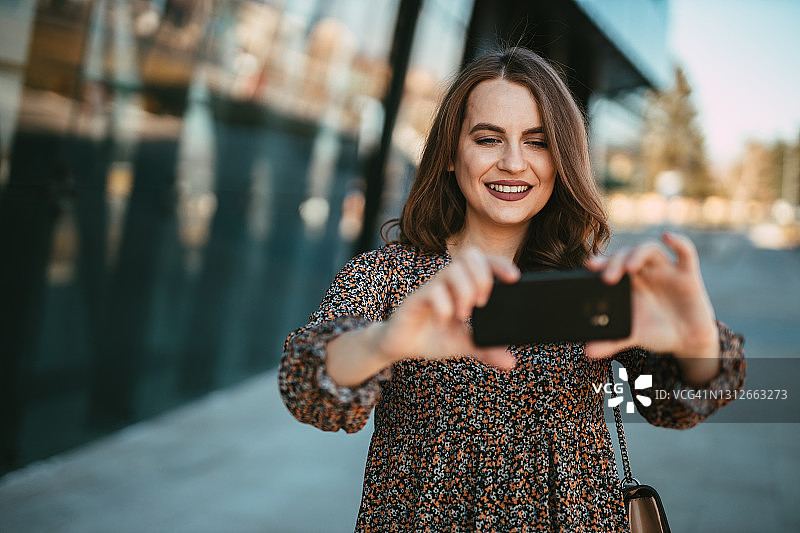 微笑的女人在城市的街道上用智能手机自拍图片素材