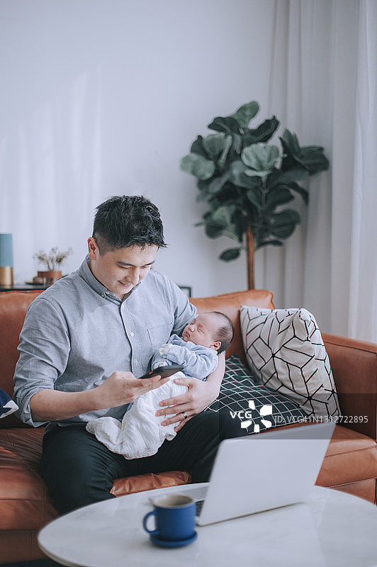 亚洲华人男子在家工作，一边用智能手机看短信，一边抱着他的小儿子坐在客厅的沙发上，用笔记本电脑图片素材