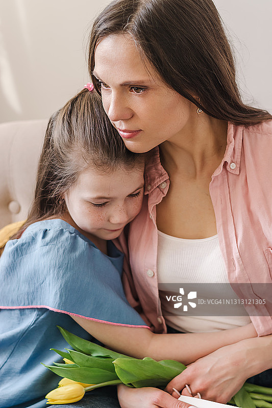 坐在家里沙发上的妈妈拥抱着她的女儿。妈妈手里拿着一张自制的贺卡和一束黄色的郁金香。母亲节、3月8日和生日的概念图片素材