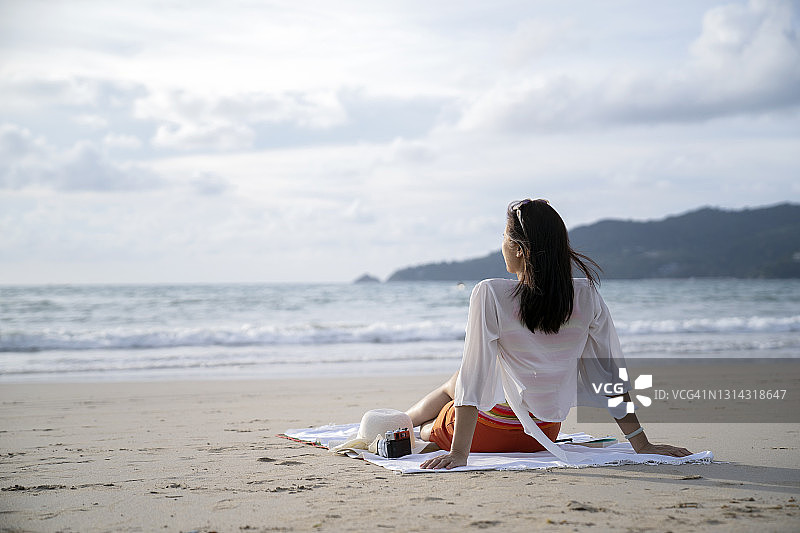 后视图有吸引力的年轻女子坐在海滩上和她看着大海。幸福和自由在大自然中。图片素材