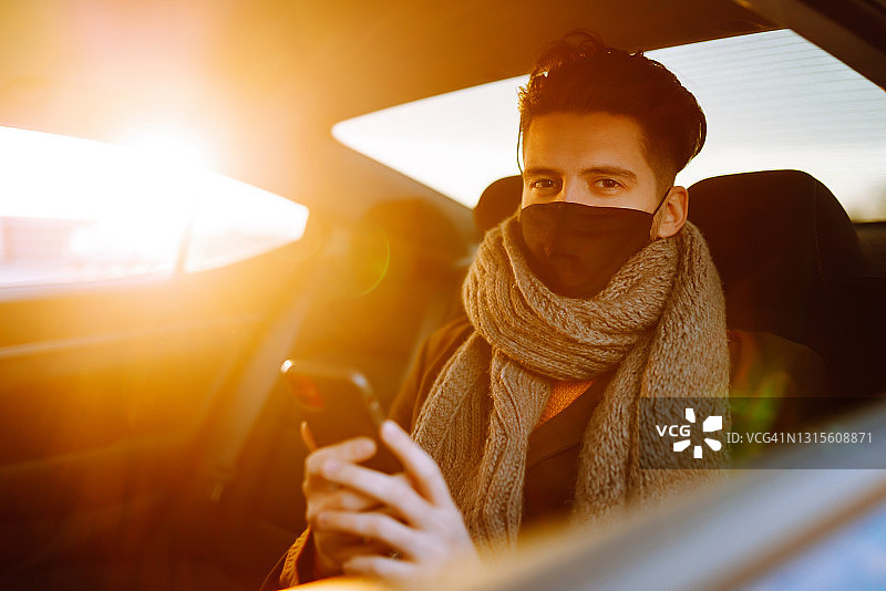 年轻男子乘客戴着防护医用口罩在出租车后座上用手机。在去办公室的路上工作。covid - 2019。图片素材
