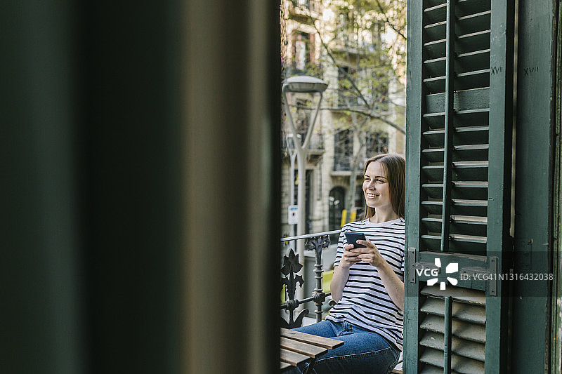 带着手机坐在度假屋阳台上做白日梦的微笑女人图片素材
