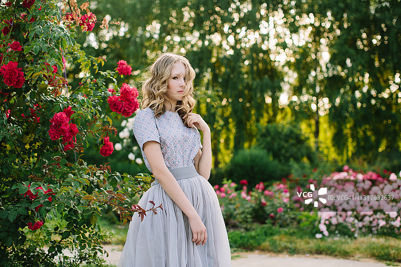 年轻的女孩穿着华丽的裙子，在夕阳下的公园里，玫瑰花丛旁图片素材