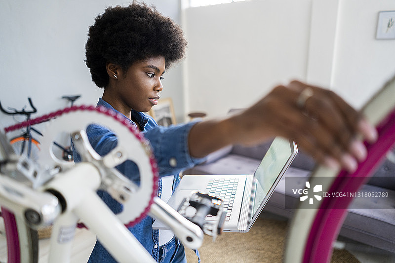 非洲发型的女人看教程在笔记本电脑上，同时修理自行车在家图片素材