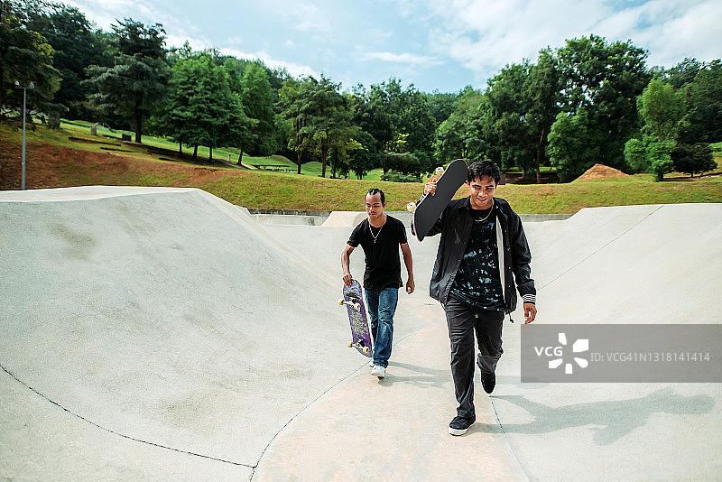 两个亚洲男性滑板手拿着他们的滑板，走向一个滑板公园图片素材
