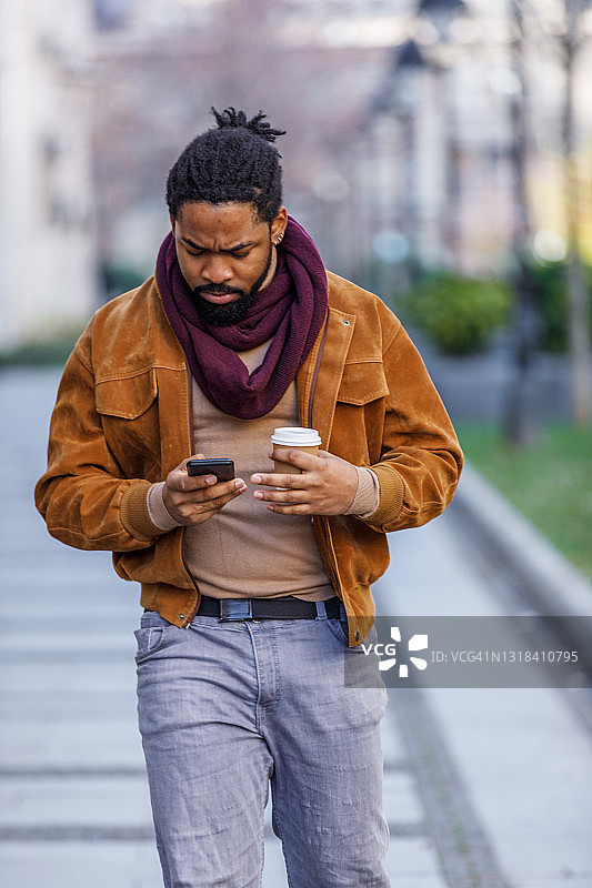 一位年轻的非洲裔美国人拿着智能手机，一边喝着咖啡，一边在市区里散步。图片素材