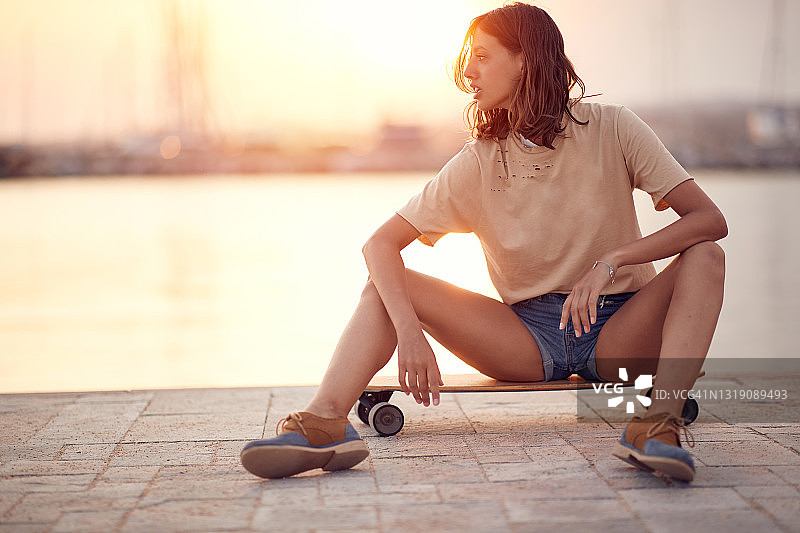 一个年轻美丽的女孩坐在海边码头上的滑板上，一边看日落，一边休息。夏天,大海,假期图片素材