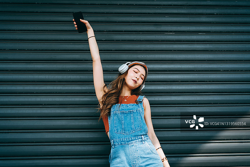 无忧无虑微笑的年轻亚洲女人拿着智能手机，在户外的彩色墙壁和阳光下，她闭着眼睛跳舞，戴着耳机听音乐。音乐和生活方式图片素材