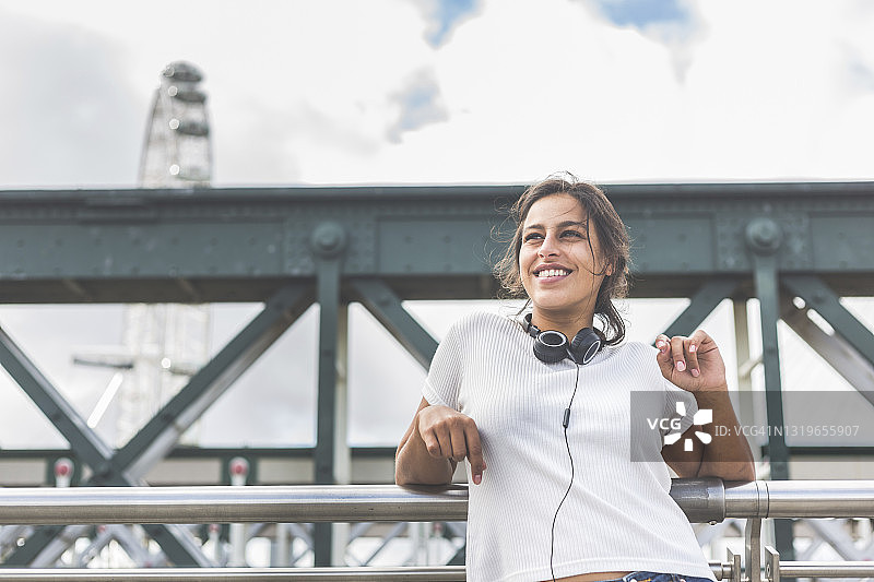 英国伦敦一座桥的栏杆上，一位面带微笑的女子戴着耳机看向别处图片素材