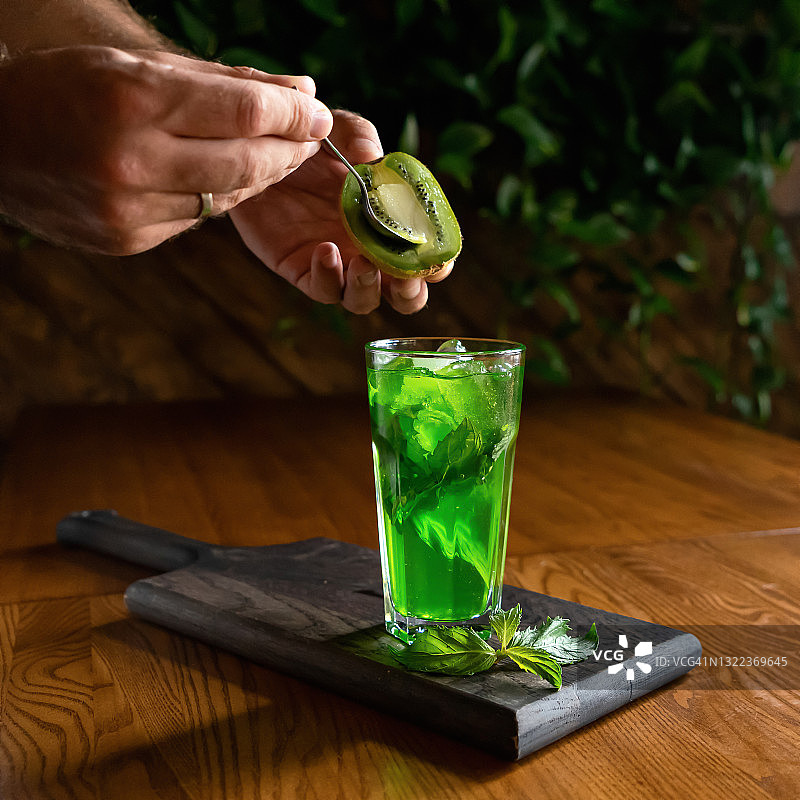 创造清爽饮料。男手用绿薄荷饮料将一茶匙猕猴桃放入玻璃杯中。健康的自然产物。大自然的明亮色彩。模糊的背景。软的焦点。本空间图片素材