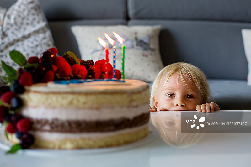 穿着蓝色衬衫的漂亮的三岁小男孩，在室内庆祝自己的生日，吹着家里烤蛋糕上的蜡烛图片素材