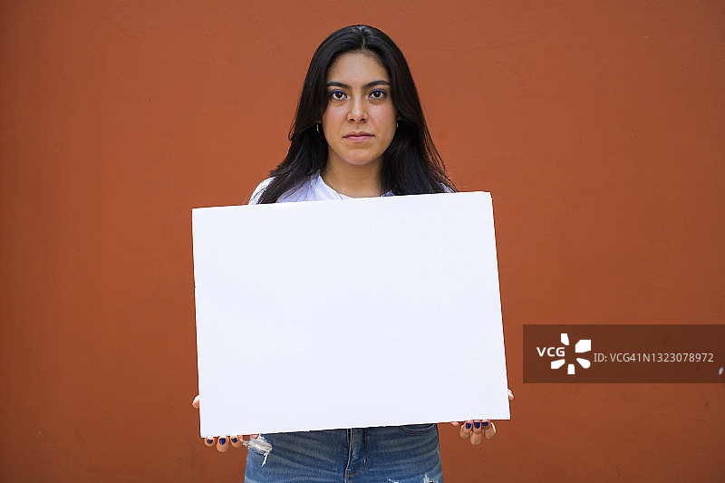 年轻的拉丁美洲妇女举着一个空白的标志，严肃的，红橙色的背景图片素材