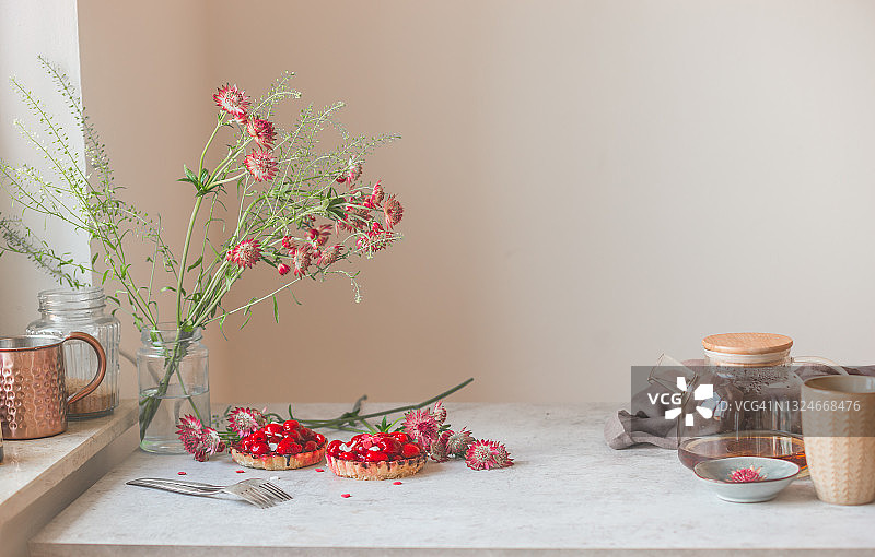 在家喝茶:自家制作的红莓果馅饼，茶罐里的茶，茶壶里的茶，浅灰色的厨房桌子上盛开的红花和叉子，墙的背景是窗台图片素材