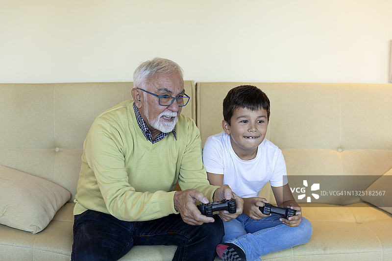 一个快乐的孩子和他的爷爷在客厅里玩电脑游戏。图片素材