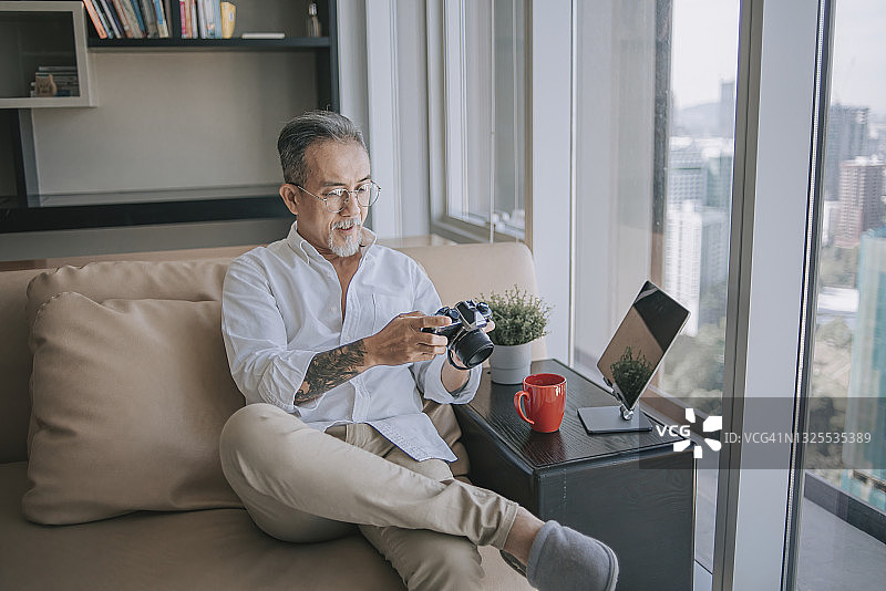 亚洲华人退休老人放松在客厅看着他的相机和数字平板电脑图片素材
