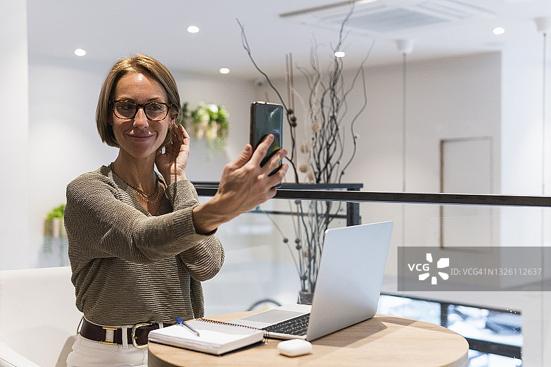 微笑的女人拿着笔记本电脑在咖啡店用智能手机自拍图片素材