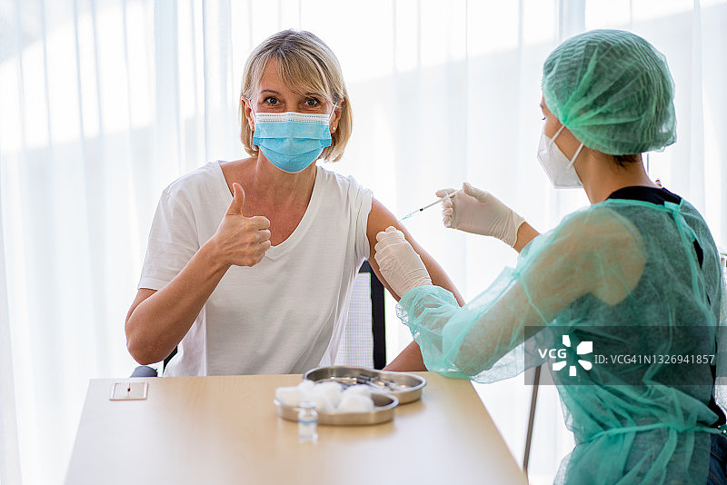 佩戴卫生口罩、身着绿色制服的护士或医务人员为年长女性注射Covid-19疫苗，竖起大拇指图片素材