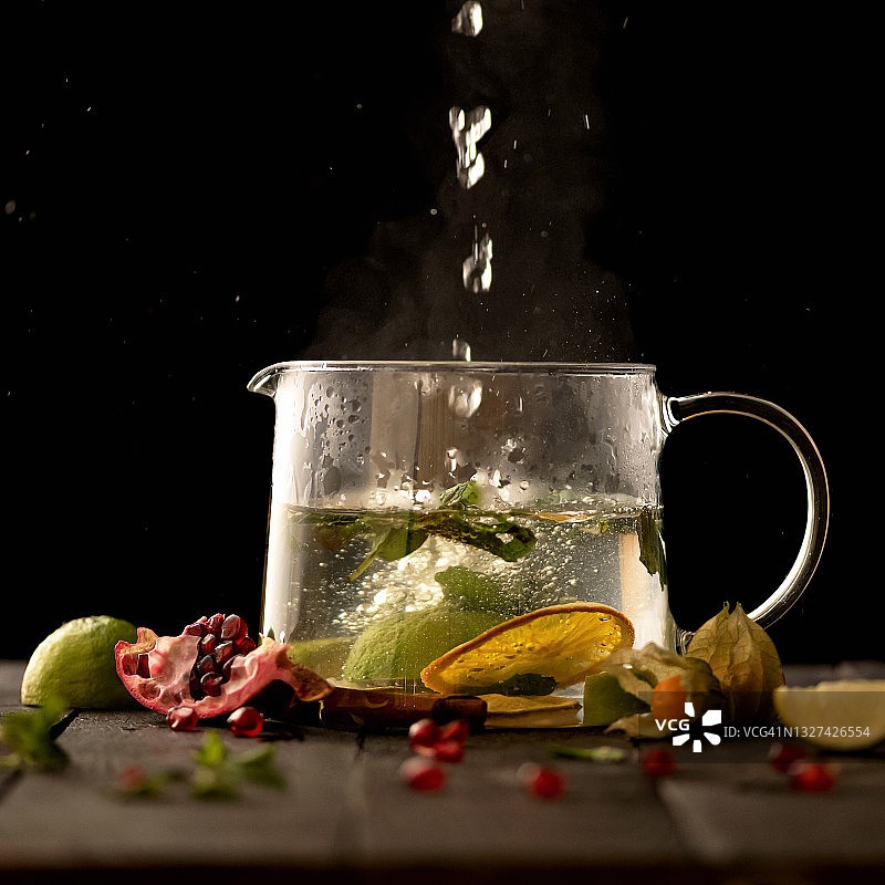 开水倒入玻璃茶壶与水果茶在黑色的背景。维生素饮料从酸橙和柠檬片，石榴籽和酸浆花。茶道的概念图片素材
