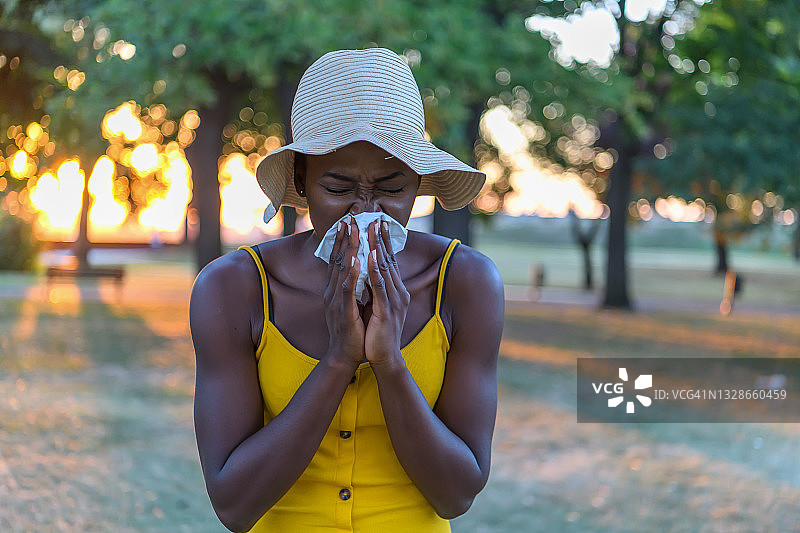 年轻的非洲妇女与感冒或流感是擤她的鼻子在公园。图片素材