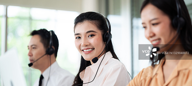 亚洲呼叫中心的工作人员戴着带有麦克风的耳机。在办公桌和电脑前服务顾客时微笑。服务理念与咨询。通信的概念。图片素材