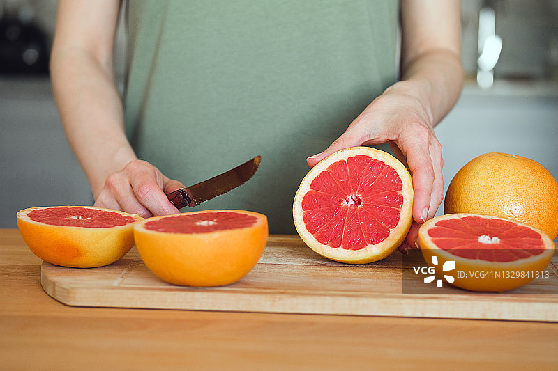 一个女孩或女人正拿着刀，在切菜板上把一个红色的成熟的葡萄柚切成两半，背景是一张木制的厨房桌子。素食、纯素食和生食的概念。图片素材