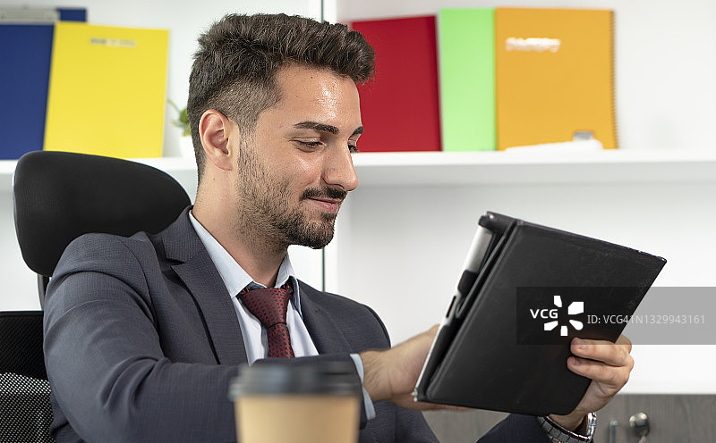 开朗的年轻职业商人，穿着西装，坐在现代办公室的办公桌前，用着电子平板电脑。图片素材