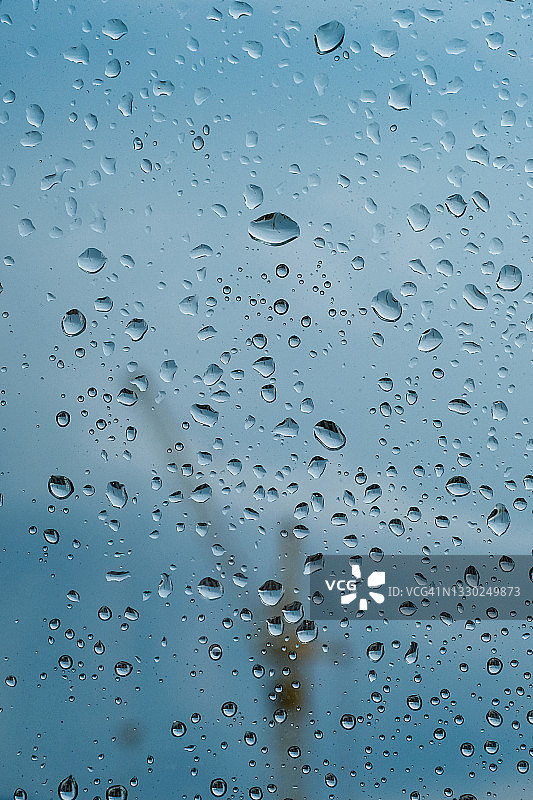 阴天，雨点落在窗户上。雨的天气，雨的背景和散景。雨后，蓝色的天空映衬着窗玻璃上的水滴。背景是一台施工起重机。图片素材