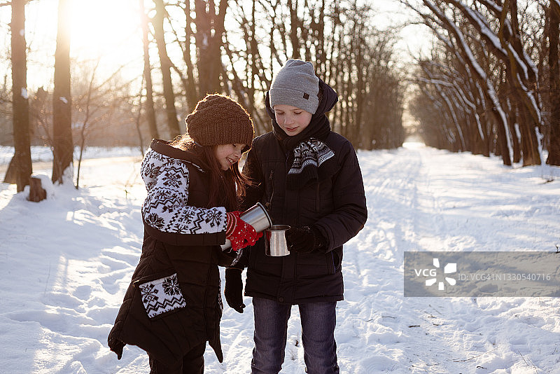 冬天，在一个白雪皑皑的公园里，一个小女孩从保温瓶里给一个男孩倒茶，倒进一个金属杯里。户外休闲的概念。图片素材