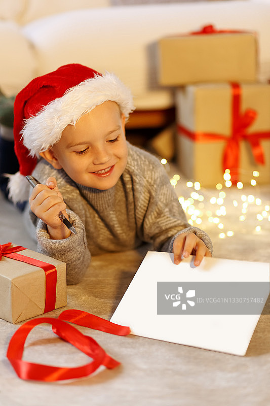 小男孩戴着圣诞帽给圣诞老人写信，在圣诞装饰附近，复制空白的空白纸，图片素材
