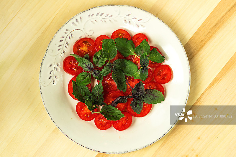 红色的番茄片和罗勒叶放在白色的盘子上，木桌背景。健康食品。图片素材