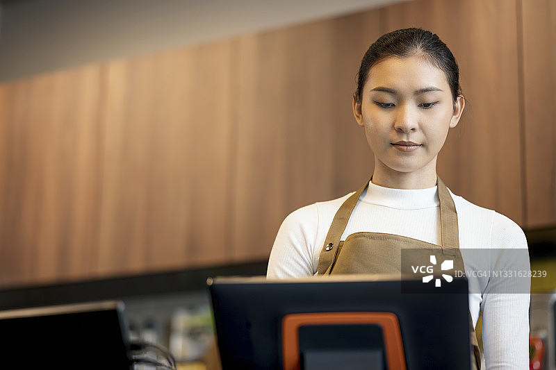 更新每日销售报表。低角度的年轻亚洲女咖啡师使用电脑更新或在线订单的咖啡馆的顾客。销售点系统和销售洞察概念。图片素材