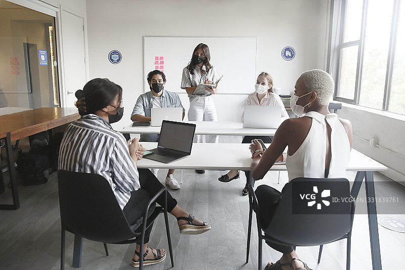 一群多种族的千禧一代，穿着时髦的休闲装，戴着防护口罩，在一间灯光明亮的会议室里，拿着笔记本电脑一起头脑风暴。图片素材