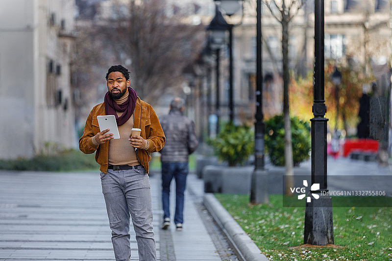 一个非裔美国年轻人在一个秋日走在城市的街道上，一边喝咖啡一边使用电子平板电脑。图片素材