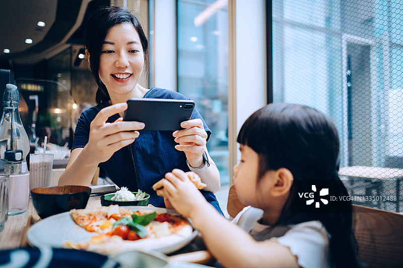 年轻的亚洲妈妈在餐厅用智能手机拍下可爱的女儿。一家人在一起享受一顿快乐的晚餐。家庭生活方式和科技图片素材