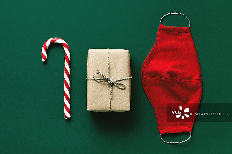 红色医用防护口罩，藤条形状的条纹糖果，绿色圣诞背景上的礼物。预防和预防COVID - 19大流行的传播。一个快乐健康的圣诞节的概念图片素材